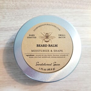 May include: A silver tin with a brown label that reads "GLORYBEENATURALS.ETSY.COM", "HAND CRAFTED", "SMALL BATCH", "BEARD BALM", "MOISTURIZE & SHAPE", "Ingredients: coconut oil, shea butter, beeswax, avocado oil, lemon, clove bud, sandalwood, nutmeg essential oil", "Sandalwood Spice", "1.75 oz (49.6 g)". The label also features a drawing of a bee.