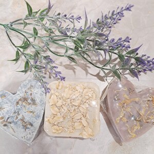 May include: Three handmade soaps in heart and square shapes. One heart-shaped soap is light blue with dried botanicals. The square soap is beige with visible oats. The other heart-shaped soap is pink with dried petals. A sprig of lavender is in the background.