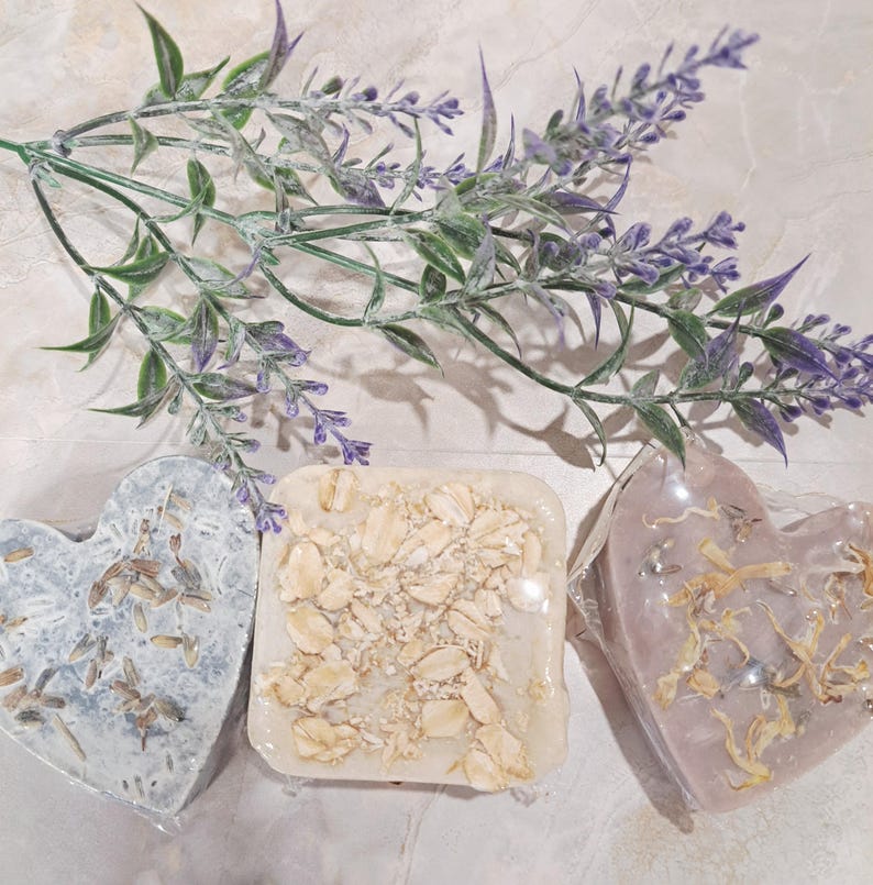 May include: Three handmade soaps in heart and square shapes. One heart-shaped soap is light blue with dried botanicals. The square soap is beige with visible oats. The other heart-shaped soap is pink with dried petals. A sprig of lavender is in the background.