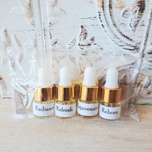 May include: Four small glass dropper bottles with gold-colored collars and white rubber bulbs, encased in a clear plastic bag. Each bottle features a white label with black text reading Radiance, Refresh, Rejuvenate, and Reborn respectively.