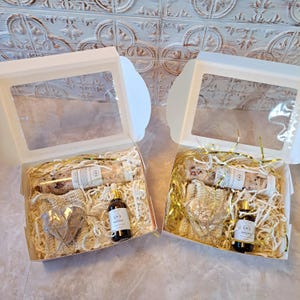 May include: Two gift boxes with clear window lids. Each box contains a glass bottle, a small bottle of oil, a heart-shaped item, and decorative filler. The boxes are on a light-colored surface.