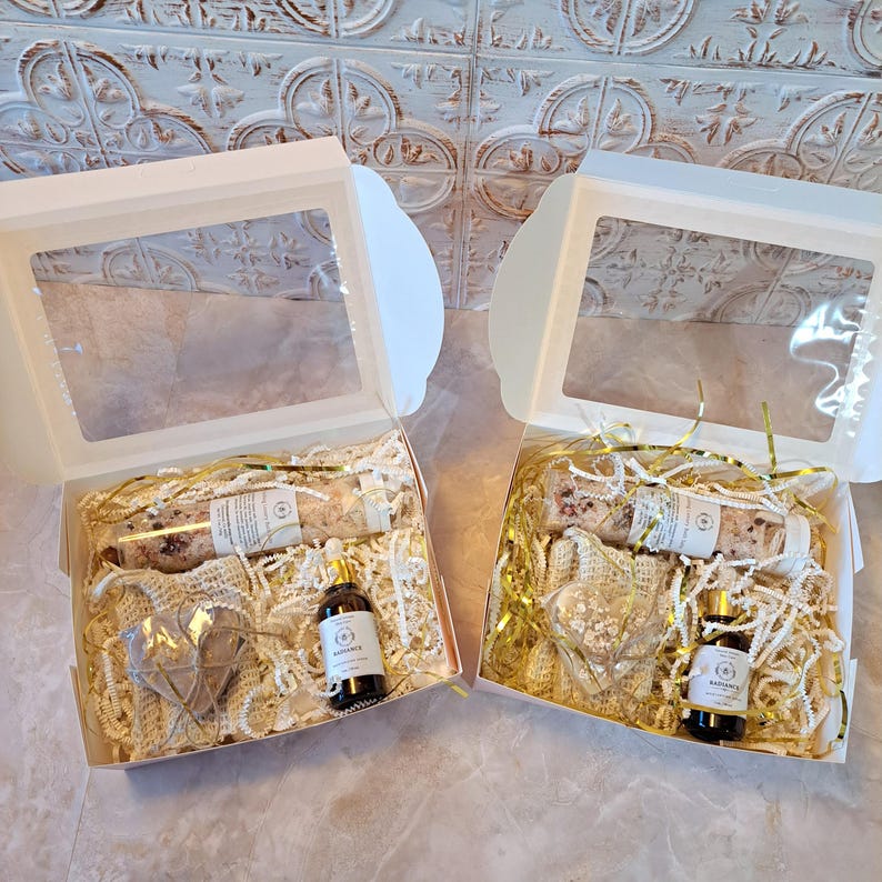 May include: Two gift boxes with clear window lids. Each box contains a glass bottle, a small bottle of oil, a heart-shaped item, and decorative filler. The boxes are on a light-colored surface.