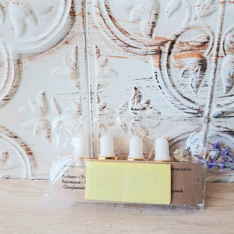 May include: Four small glass bottles with white dropper caps are arranged in a row inside a clear plastic sleeve. A brown paper label with printed text is attached to the front, partially covered by a rectangular yellow paper insert.