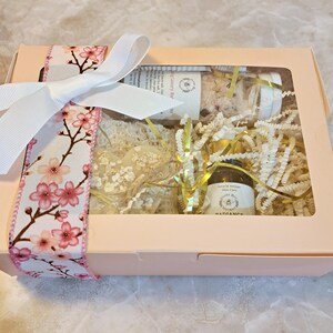 May include: A light pink gift box tied with a white bow and a floral ribbon featuring pink cherry blossoms. The box contains bath products, including a jar of bath salts, a small bottle of oil, and a bar of soap, all nestled in decorative paper.