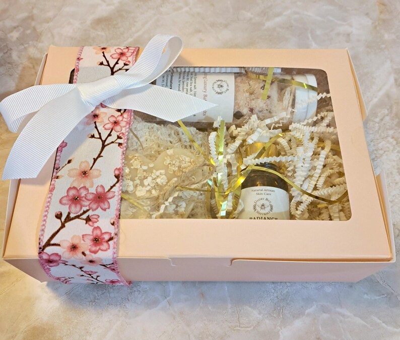 May include: A light pink gift box tied with a white bow and a floral ribbon featuring pink cherry blossoms. The box contains bath products, including a jar of bath salts, a small bottle of oil, and a bar of soap, all nestled in decorative paper.