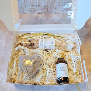 May include: A white gift box filled with self-care items. Includes a bottle of moisturizing serum, a heart-shaped soap, bath salts, and a small wrapped item. The box is decorated with gold ribbon and white paper filler.