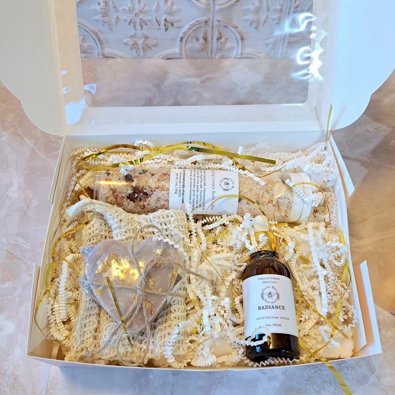 May include: A white gift box filled with self-care items. Includes a bottle of moisturizing serum, a heart-shaped soap, bath salts, and a small wrapped item. The box is decorated with gold ribbon and white paper filler.