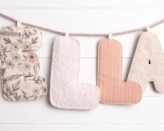May include: A banner spelling "ELLA" with fabric letters. The "E" is patterned with a floral design, the "L" is fluffy white, the second "L" is pink with vertical stripes, and the "A" is a neutral linen color.