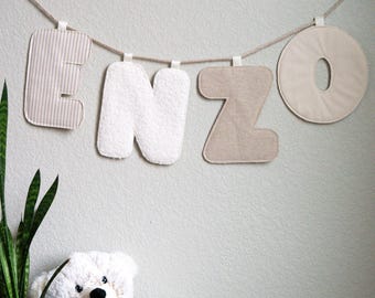May include: A banner spelling "ENZO" hangs against a light gray wall. The letters are in various textures and colors: striped, white terry cloth, and tan. A plush white teddy bear sits below, next to a potted plant.