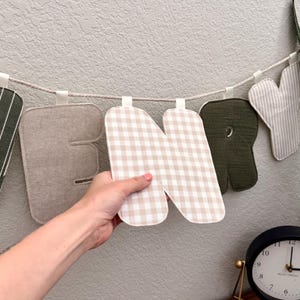 May include: A fabric letter garland featuring individual stuffed letters suspended on a rope. The letters display various patterns including beige gingham, solid olive green, and striped fabric. One letter N is held up to show the texture.