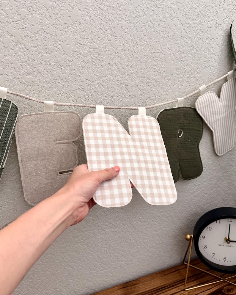 May include: A fabric letter garland featuring individual stuffed letters suspended on a rope. The letters display various patterns including beige gingham, solid olive green, and striped fabric. One letter N is held up to show the texture.