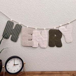 May include: A fabric banner spelling the name HENRY, featuring individual letters in alternating patterns of dark green stripes, solid beige, beige gingham, solid dark green, and beige stripes, all suspended from a single white cord.