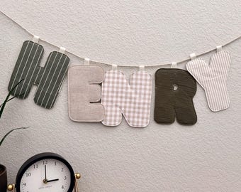 May include: A banner spelling "HENRY" with fabric letters in various patterns, including stripes and gingham, hangs against a textured wall. A black analog clock with gold accents sits on a wooden surface below.