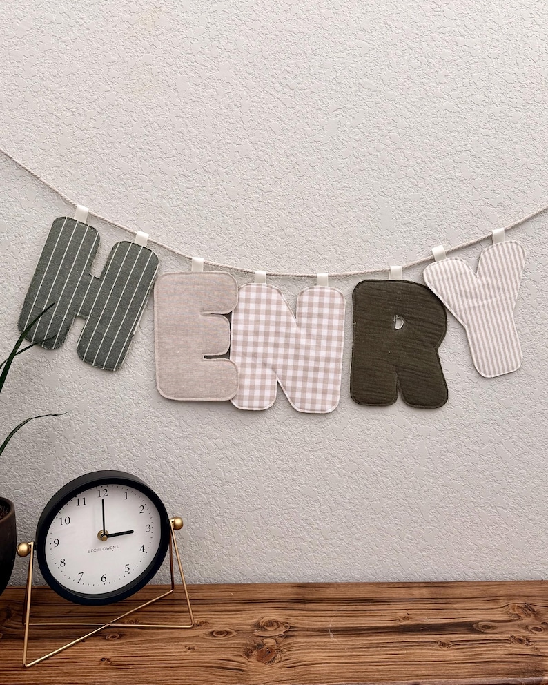 May include: A fabric banner spelling the name HENRY, featuring individual letters in alternating patterns of dark green stripes, solid beige, beige gingham, solid dark green, and beige stripes, all suspended from a single white cord.