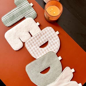 May include: A set of five fabric letters spelling the word "TODAY" in various textures and patterns. The letters feature sage green plaid, white sherpa, beige gingham, solid sage green, and cream corduroy, each with a small white fabric tag.