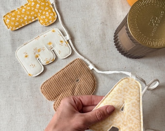May include: A fabric banner featuring four stuffed, padded letters spelling the word TEA. The letters are crafted from various textiles, including yellow floral print, cream floral print, and tan corduroy, all strung together on a white cord.