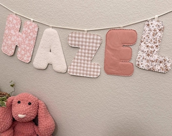 May include: A decorative banner spelling "HAZEL" in fabric letters, each with a unique pattern: floral, textured white, gingham, corduroy, and floral. A pink plush bunny sits below the banner on a wooden surface.
