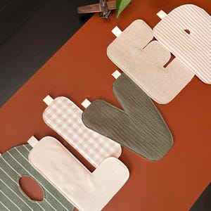 May include: A set of fabric letters spelling the word LOVE, featuring various textures and patterns including olive green stripes, beige gingham, solid olive green, and beige stripes. Each letter has a small white fabric loop attached to the top edge.