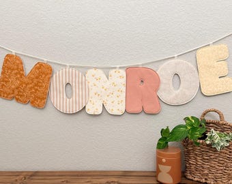 May include: A banner spelling "MONROE" with fabric letters in various patterns and colors, including floral, striped, and solid textures. The banner hangs above a wooden shelf with a decorative jar and a woven basket.