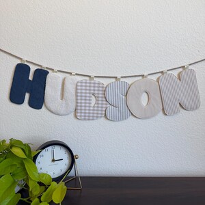 May include: A fabric banner spelling the name HUDSON, featuring individual letters in navy blue, cream, beige gingham, and grey striped patterns, all suspended from a single length of twine with small fabric loops at the top of each letter.