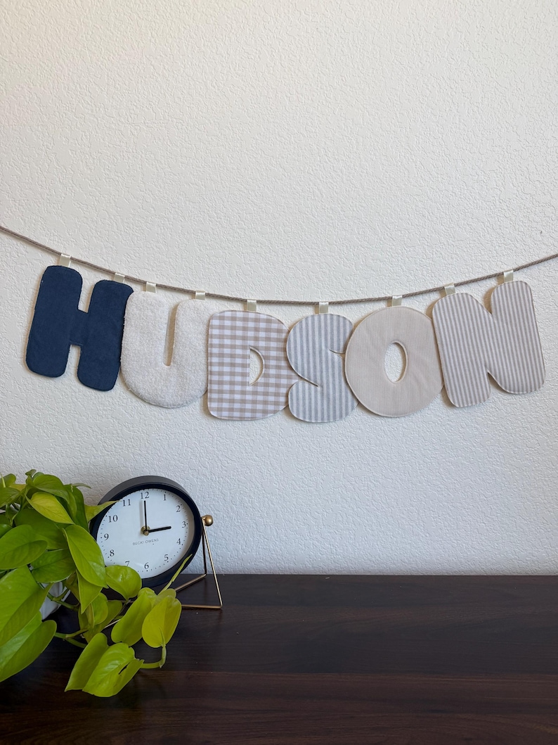 May include: A fabric banner spelling the name HUDSON, featuring individual letters in navy blue, cream, beige gingham, and grey striped patterns, all suspended from a single length of twine with small fabric loops at the top of each letter.