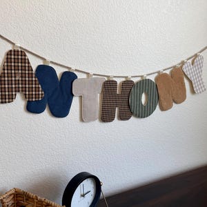 May include: A fabric letter banner spelling the name ANTHONY, featuring individual letters in various textures and patterns including brown plaid, solid navy blue, beige, green with white stripes, and tan corduroy, all suspended on a rope.