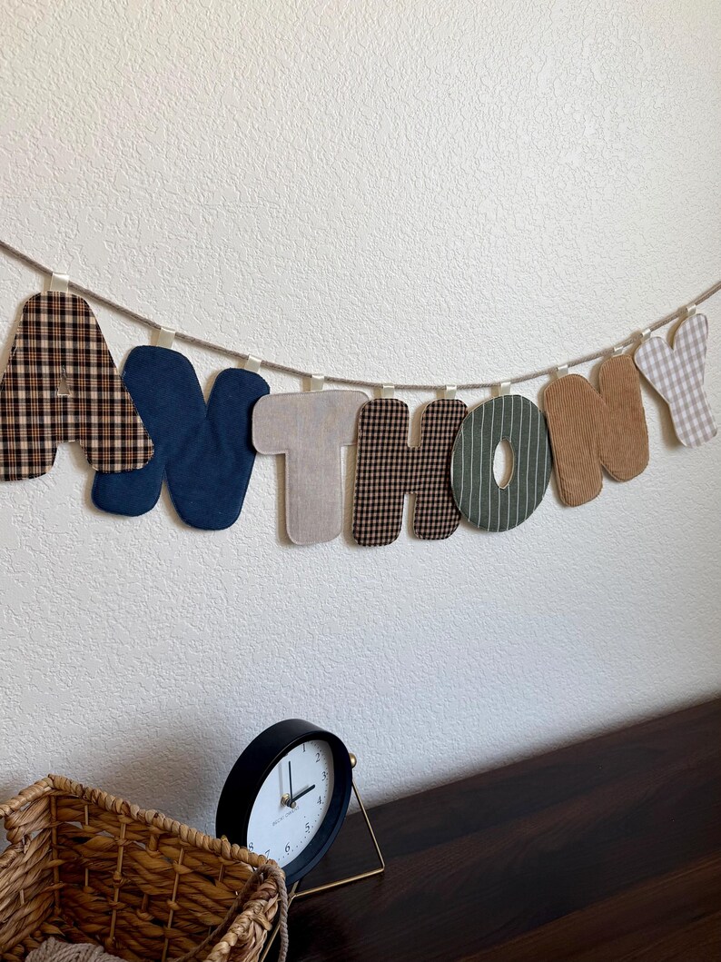 May include: A fabric letter banner spelling the name ANTHONY, featuring individual letters in various textures and patterns including brown plaid, solid navy blue, beige, green with white stripes, and tan corduroy, all suspended on a rope.