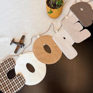 May include: A decorative fabric banner spelling the name COOPER, featuring individual letters crafted from various textiles including brown and white plaid, solid cream, tan corduroy, beige stripes, and solid beige, all strung on a thick rope.