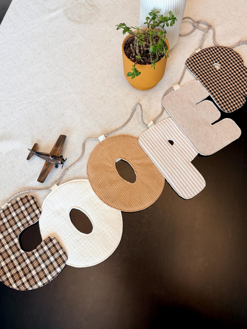 May include: A decorative fabric banner spelling the name COOPER, featuring individual letters crafted from various textiles including brown and white plaid, solid cream, tan corduroy, beige stripes, and solid beige, all strung on a thick rope.