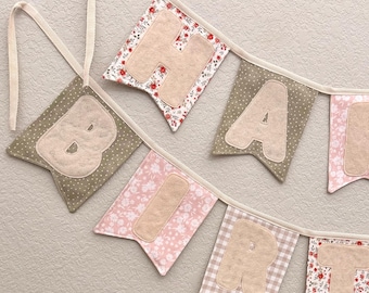 May include: A handmade banner spelling out "BIRTH". Each flag features a different patterned fabric, including floral, polka dot, and gingham. The letters are a soft beige color, and the banner is strung on a cream-colored ribbon.