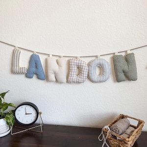 Puede incluir: Una guirnalda de tela que deletrea el nombre LANDON con letras acolchadas individuales colgadas de una cuerda. Las letras presentan varios patrones y colores, incluyendo rayas grises, azul sólido, crema sólido, cuadros vichy beige y verde salvia sólido.