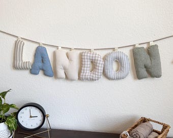 May include: A fabric banner spelling the name LANDON with individual stuffed letters hanging from a string. The letters feature various patterns and colors, including grey stripes, solid blue, solid cream, beige gingham, and solid sage green.
