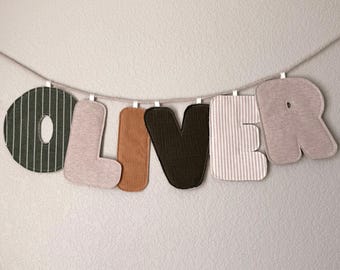 May include: A banner spelling "OLIVER" with fabric letters. The letters are in various colors and patterns, including green and white stripes, tan, brown corduroy, and gray. The letters are suspended from a string.