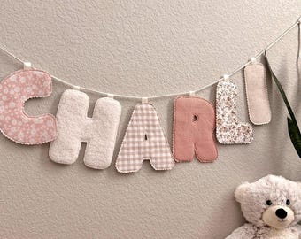 May include: A decorative banner spelling "CHARLI" with fabric letters in various patterns and textures, including floral, gingham, and ribbed designs. The banner is strung on a white cord and displayed against a neutral wall. A plush teddy bear sits below.