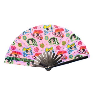 May include: Pink hand fan featuring a repeating pattern of cartoon characters with red, yellow, and green hair, and blue eyes. The fan also has colorful flower designs and the text "JUSTONLYFANS". The fan has a black wooden frame.