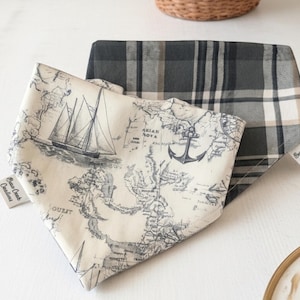 May include: Two dog bandanas. One features a nautical map design with a sailboat and anchor in navy blue on an off-white background. The other is a gray and white plaid pattern. Both have a tag that says "Bear Creek Creations."