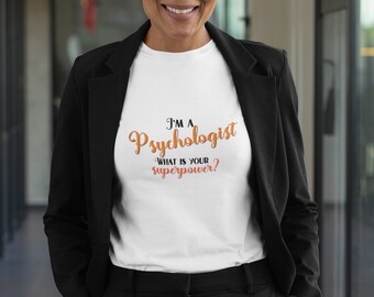 May include: A woman wearing a white t-shirt with the text "I'm a Psychologist What is your superpower?" in orange lettering. She is wearing a black blazer and black pants.