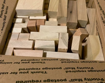 Op de afbeelding: Een kartonnen doos gevuld met verschillende maten en vormen van onbewerkte houten blokken. De doos is gelabeld met de tekst "Priority Mail" en "postage required".