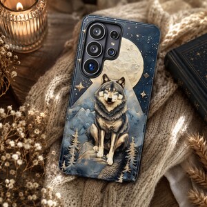 May include: A rectangular smartphone case featuring a dark blue night sky background with a large full moon, golden stars, and a central illustration of a grey and white wolf sitting on a rocky mountain peak surrounded by pine trees.