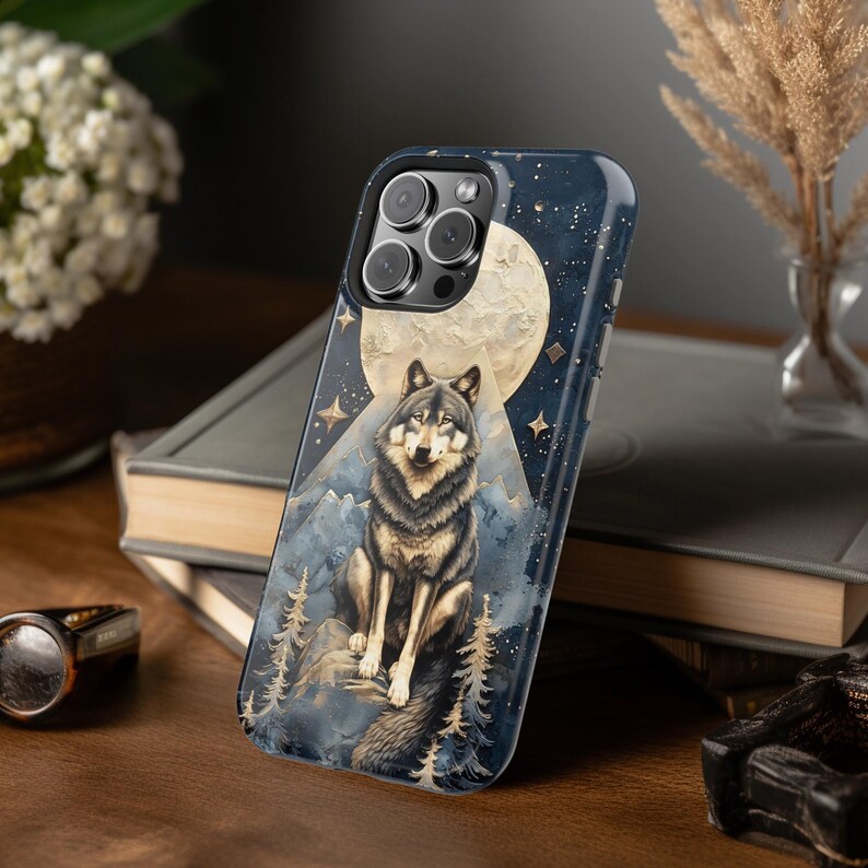 May include: A smartphone case featuring a dark blue night sky background with a large full moon, a central illustration of a grey and brown wolf sitting on a rock, and stylized mountain and pine tree silhouettes with gold star accents.
