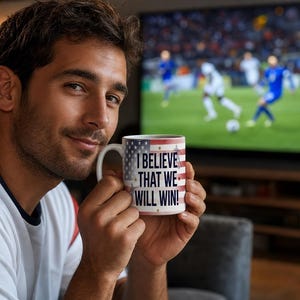 May include: A white ceramic mug with a patriotic American flag design and the text "I BELIEVE THAT WE WILL WIN!" is held up in front of a television screen. The mug has a white handle and a red, white, and blue design.