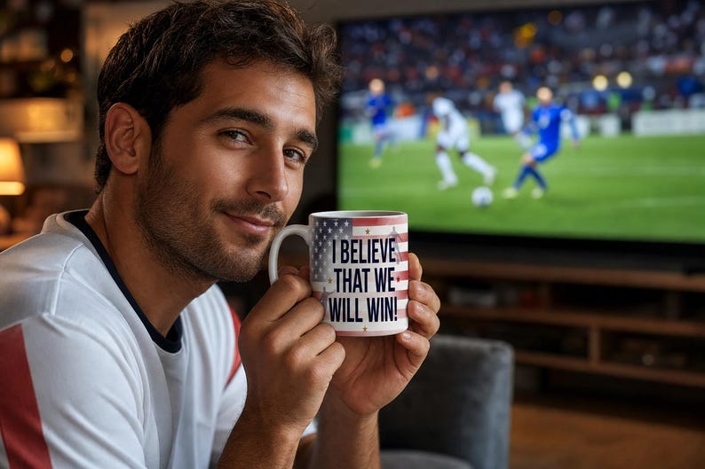 May include: A white ceramic mug with a patriotic American flag design and the text "I BELIEVE THAT WE WILL WIN!" is held up in front of a television screen. The mug has a white handle and a red, white, and blue design.