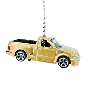 May include: A gold-colored toy pickup truck with black wheels and a black bed. The truck is suspended from a silver chain, likely a ceiling fan pull. The truck has a detailed design, including headlights and taillights.
