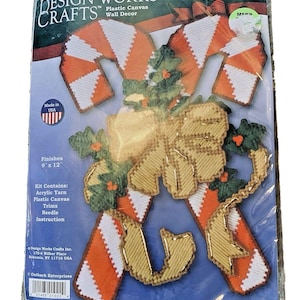 May include: A Design Works Crafts plastic canvas wall decor kit. The kit includes acrylic yarn, plastic canvas, trims, and a needle. The finished product measures 9 x 12 inches. The design features candy canes and a bow with green holly accents.
