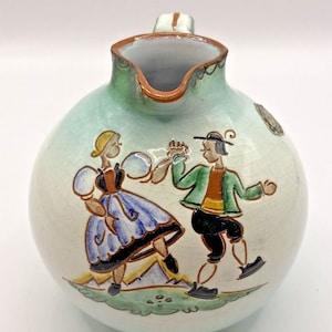 May include: A white ceramic pitcher with a green glaze and a brown handle. The pitcher features a hand-painted scene of a man and woman in traditional folk costumes dancing. The scene is set against a green and yellow background.