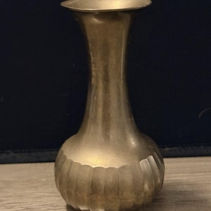 May include: A vintage brass vase with a fluted lower body and a slender, curved neck. The vase has a warm, metallic gold color and a flared rim. The vase is approximately 7 inches tall.