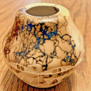 May include: A small, beige ceramic vase with a unique design. The vase features a round body with a narrow opening and a decorative pattern of blue, black, and red lines. The bottom has a geometric design.