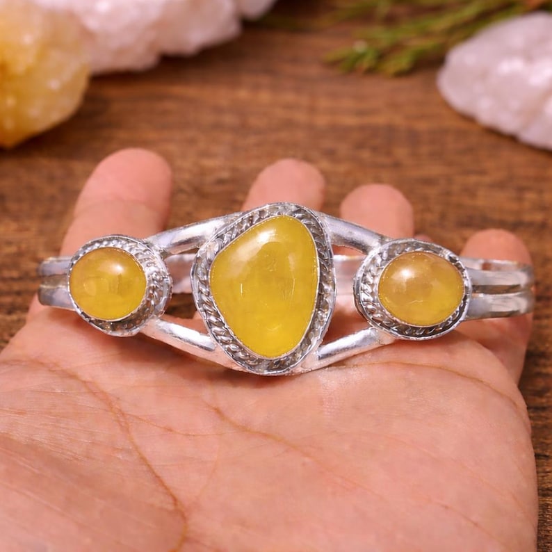 May include: A silver cuff bracelet featuring three large, oval-shaped yellow gemstones. The central stone is larger and more irregularly shaped than the two flanking stones. The bracelet is held in a hand, showcasing the jewelry's design and color.
