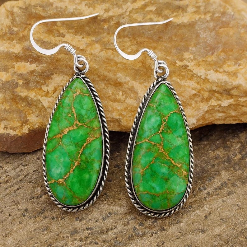 May include: A pair of silver-toned earrings with teardrop-shaped green stones. The stones have gold-colored veining and are framed by a twisted silver rope design. The earrings have a simple hook closure.