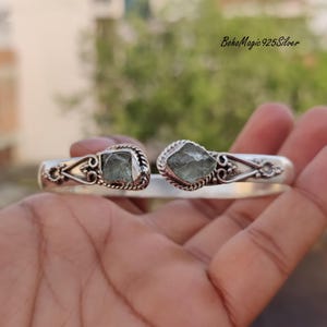 Puede incluir: Un brazalete de plata con dos piedras de aguamarina verde engastadas en el centro. El brazalete es abierto y ajustable.